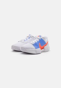 Et par hvide sneakers med blå og orange detaljer, teksturerede såler og hvide snørebånd på en ensfarvet hvid baggrund.