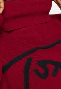Czerwony sweter robiony na drutach z wysokim kołnierzem, posiadający wyrazisty czarny wzór graficzny. Tekstura jest gładka i prążkowana.