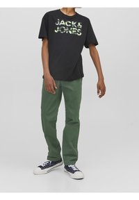 Czarna koszulka z grafiką "JACK & JONES" w kolorze zielonym, zestawiona z zielonymi spodniami i granatowymi sneakersami z białymi podeszwami.