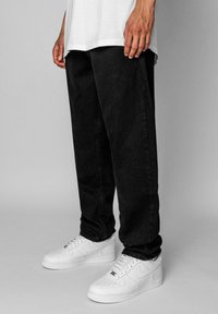 Sort denimbukser med en afslappet pasform, båret med hvide sneakers. Stoffet har en glat tekstur og subtile syninger.