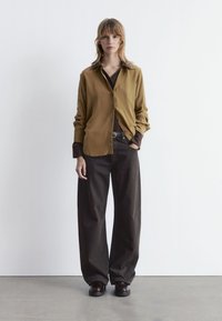 Long-sleeve, button-up shirt in mustard with a dark brown collar paired with wide, dark brown trousers and glossy shoes. Simple design.