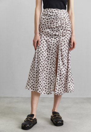 Kvinde iført en hvid midinederdel med sort blomsterprint, front rynker og slids, kombineret med sorte slip-on træsko med guldkæde detalje.