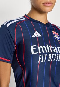 Pogled od blizu na osebo, ki nosi športno majico mornarsko modre barve z rdečimi in modrimi navpičnimi črtami, belim logotipom Adidas in besedilom "Emirates Fly Better".