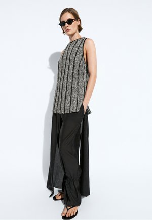 Donna con occhiali da sole neri, top senza maniche nero e bianco a fantasia, pantaloni neri larghi con lunghi pannelli laterali e sandali neri.