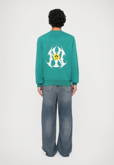 Felpa turchese con un grande grafico bianco e un design di faccia gialla sul retro, abbinata a jeans in denim azzurro a vestibilità larga.