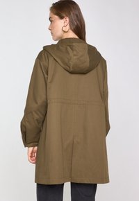 Frau mit braunen Haaren, die große Creolen trägt und einen olivgrünen Kapuzenmantel anhat, von hinten vor einem schlichten Hintergrund gezeigt.