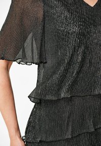 Schwarzes, plissiertes Kleid mit Rüschensleeves und Saum. Glänzender, strukturierter Stoff erzeugt vertikale Streifen. Der V-Ausschnitt betont die Struktur.