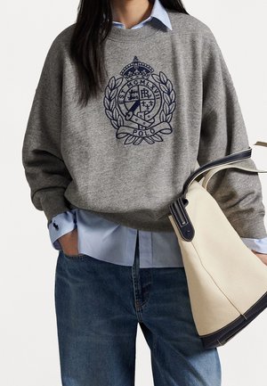 Osoba ubrana w szarą bluzę z haftowanym herbem, jasnoniebieską koszulę, niebieskie dżinsy, trzymająca beżowo-granatową torebkę na białym tle.