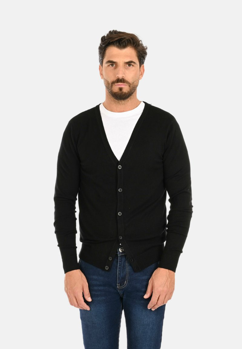 Cardigan nero lavorato a maglia con scollatura a V, bottoni monopetto e polsini a coste. Indossato sopra una maglietta bianca con jeans blu.