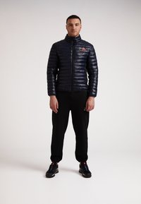 Marineblå pufferjakke med høj krave og lynlås, prydet med et lille logo. Kombinationen med sorte sweatpants og sorte sportsko.