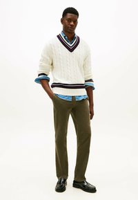 Uomo in piedi con le mani in tasca, che indossa un maglione bianco a trecce sopra una camicia blu, pantaloni oliva e mocassini neri su sfondo bianco.