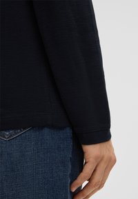 Chemise à manches longues navy avec un tissu texturé, un ourlet arrondi et des coutures plates, portée avec un jean en denim. Main reposant sur le côté.