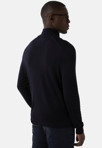 Mężczyzna w czarnym swetrze z golfem i ciemnych spodniach, pokazany od tyłu, spoglądający w bok na jednolitym tle.