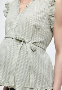 Αμάνικη μπλούζα με πράσινο-ανοιχτό πράσινο και άσπρο καρό μοτίβο gingham, με κουμπωτό μπροστινό μέρος, πιέτες και ζώνη στη μέση δεμένη σε φιόγκο σε άτομο.