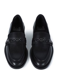 Mocasines de cuero negro con punta redondeada, que cuentan con una correa decorativa con acentos metálicos y una superficie lisa y texturizada.