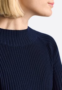 Femme blonde portant un pull côtelé bleu marine avec un col montant, montrant le profil latéral de son cou et de son visage sur un fond blanc.