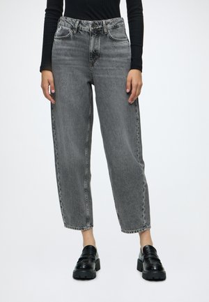 Vrouw draagt hooggetailleerde vervaagde grijze jeans met taps toelopende pasvorm en zwarte plateau loafers, staand tegen een effen achtergrond.
