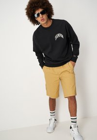 Sudadera negra con el logo "Champion", pantalones cortos amarillos, zapatillas blancas y gafas de sol negras. Tejido texturizado con puños acanalados y calcetines a rayas.