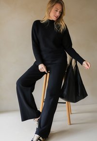 Zwarte trui en wijde broek, beide van een zachte stof. De outfit heeft een hoge halslijn en geribbelde manchetten, aangevuld met een zwarte tas.