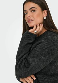 Frau mit langen braunen Haaren, die einen dunkelgrauen Strickpullover und silberne strukturierte Ohrringe und Ringe trägt, posiert mit der Hand nahe am Gesicht.