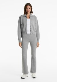 Vrouw in een grijze zip-up jack, bijpassende grijze broek, wit tanktop en witte sneakers, staand tegen een witte achtergrond.