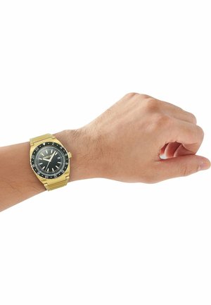 Polso maschile con orologio da polso in metallo dorato con quadrante nero e ghiera rotante a 24 ore su sfondo bianco.