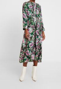 Vestido midi floral en verde oscuro, con patrones de rosas en rosa y blanco, mangas largas, diseño plisado y cuello redondo. Llevado con botas de tobillo blancas.