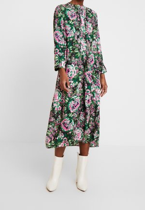 Robe midi florale en vert foncé, avec des motifs de roses roses et blanches, des manches longues, un design plissé et un col rond. Portée avec des bottines blanches.