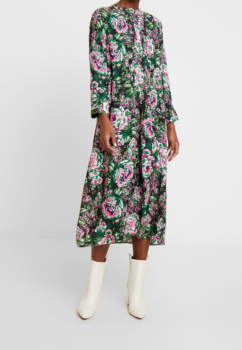 Vestido midi floral en verde oscuro, con patrones de rosas en rosa y blanco, mangas largas, diseño plisado y cuello redondo. Llevado con botas de tobillo blancas.