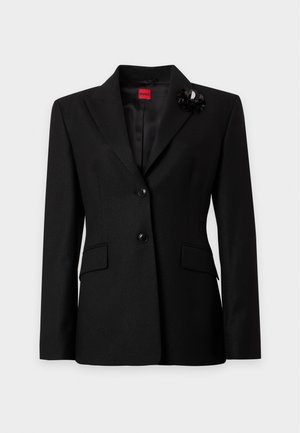 Femme debout portant un blazer noir, une chemise noire, un pantalon évasé noir et des chaussures pointues noires sur un fond clair uni.