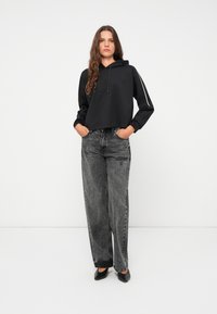 Svart cropped hettegenser med snøring, kombinert med høytlivs grå jeans. Modellen står med hendene i lommene og har på seg svarte sko.