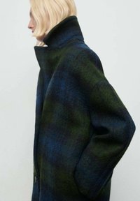 Cappotto in misto lana a scacchi blu scuro e verde, con colletto alto e tasche laterali, dall'aspetto morbido e strutturato.