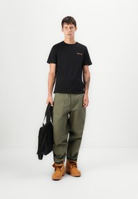 Zwarte katoenen t-shirt met een klein oranje logo, groene losse cargo broek, bruine laarzen en een zwarte tote bag.