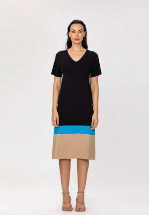 Femme debout portant une robe noire mi-longue à manches courtes, avec un bloc de couleur bleu et beige à l'ourlet, et des sandales beiges.