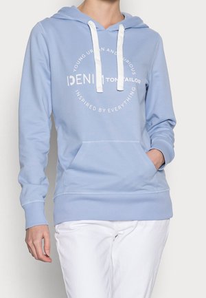 Persoon draagt een lichtblauwe hoodie met witte trekkoorden en een voorzak, witte broek, tekst "DENIM TOM TAILOR" op de voorkant van de hoodie.