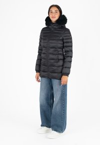Giacca piumino nera con cappuccio in pelliccia sintetica, design trapuntato, zip completa e polsini elasticizzati, abbinata a jeans a gamba larga e scarpe da ginnastica bianche.