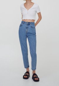 Top court blanc à côtes avec des manches courtes et trois boutons. Jean taille haute bleu clair avec une coupe fuselée et des poches avant. Sandales noires.