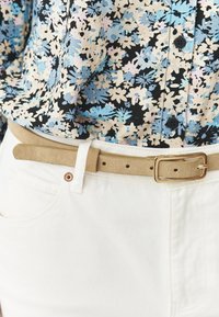 Beige suède riem met een rechthoekige gouden gesp, gedragen over witte jeans en een bloempatroon blouse in blauw, zwart en crèmekleuren.