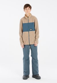 Chaqueta de forro polar en beige con un panel en azul verdoso, cierre de cremallera y parche con logo; combinada con pantalones azules y botas negras. Superficies suaves y texturizadas.