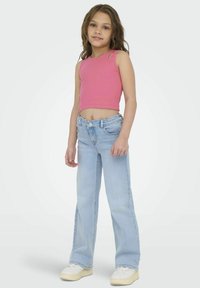 Débardeur court à côtes roses avec découpes aux épaules, associé à un jean large bleu clair et des baskets blanches, se tenant devant un fond uni.