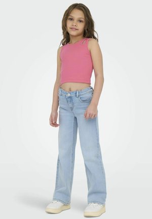 Débardeur court à côtes roses avec découpes aux épaules, associé à un jean large bleu clair et des baskets blanches, se tenant devant un fond uni.