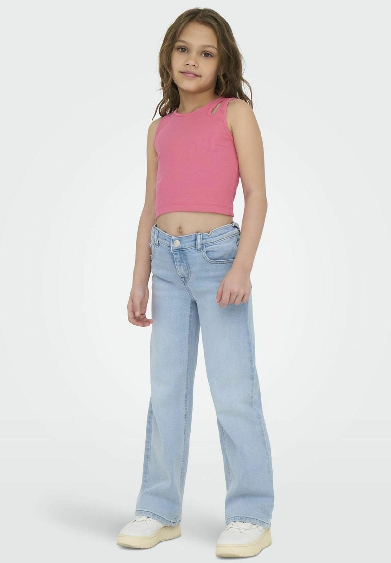 Débardeur court à côtes roses avec découpes aux épaules, associé à un jean large bleu clair et des baskets blanches, se tenant devant un fond uni.