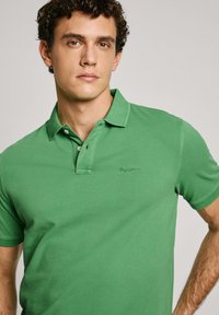 Green polo shirt made of cotton, featuring a three-button placket and embroidered logo on the left chest. Short sleeves, smooth texture.