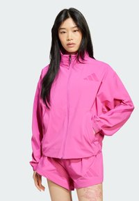 Cazadora de viento Adidas de color rosa brillante con cierre de cremallera, puños elásticos y pantalones cortos a juego. Presenta detalles con logos y un ajuste relajado.