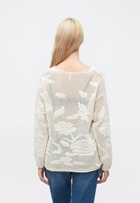 Pull tricoté crème, avec un motif en maille ouverte et des motifs floraux. Le dos présente un col rond dégagé et des manches amples.