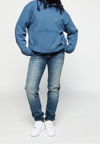 Blue sweatshirt with kangaroo pocket, paired with faded blue jeans and white sneakers. Simple design, relaxed fit, and soft texture.