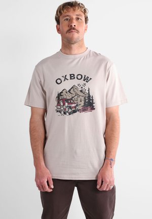 Homme portant un t-shirt beige avec le texte "OXBOW", une scène de montagne avec des arbres, un véhicule rouge et deux snowboarders avec leur équipement.