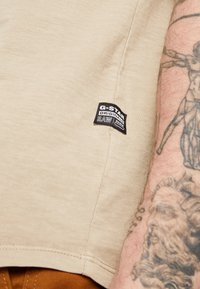 Beige katoenen t-shirt met een zwart G-Star RAW-label linksonder. De stof heeft een soepele textuur met minimale zichtbare naden.