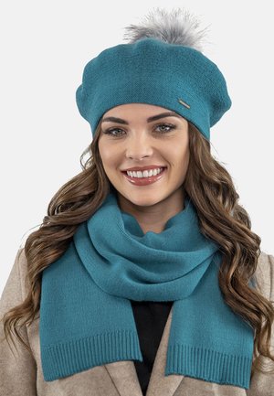 Berretto lavorato a maglia turchese con pompon in pelliccia grigia, abbinato a un foulard coordinato con bordi a coste per texture e calore.