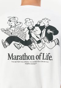 White t-shirt featuring a black graphic illustration of three cartoon figures walking. Text reads "Marathon of Life" below the image.